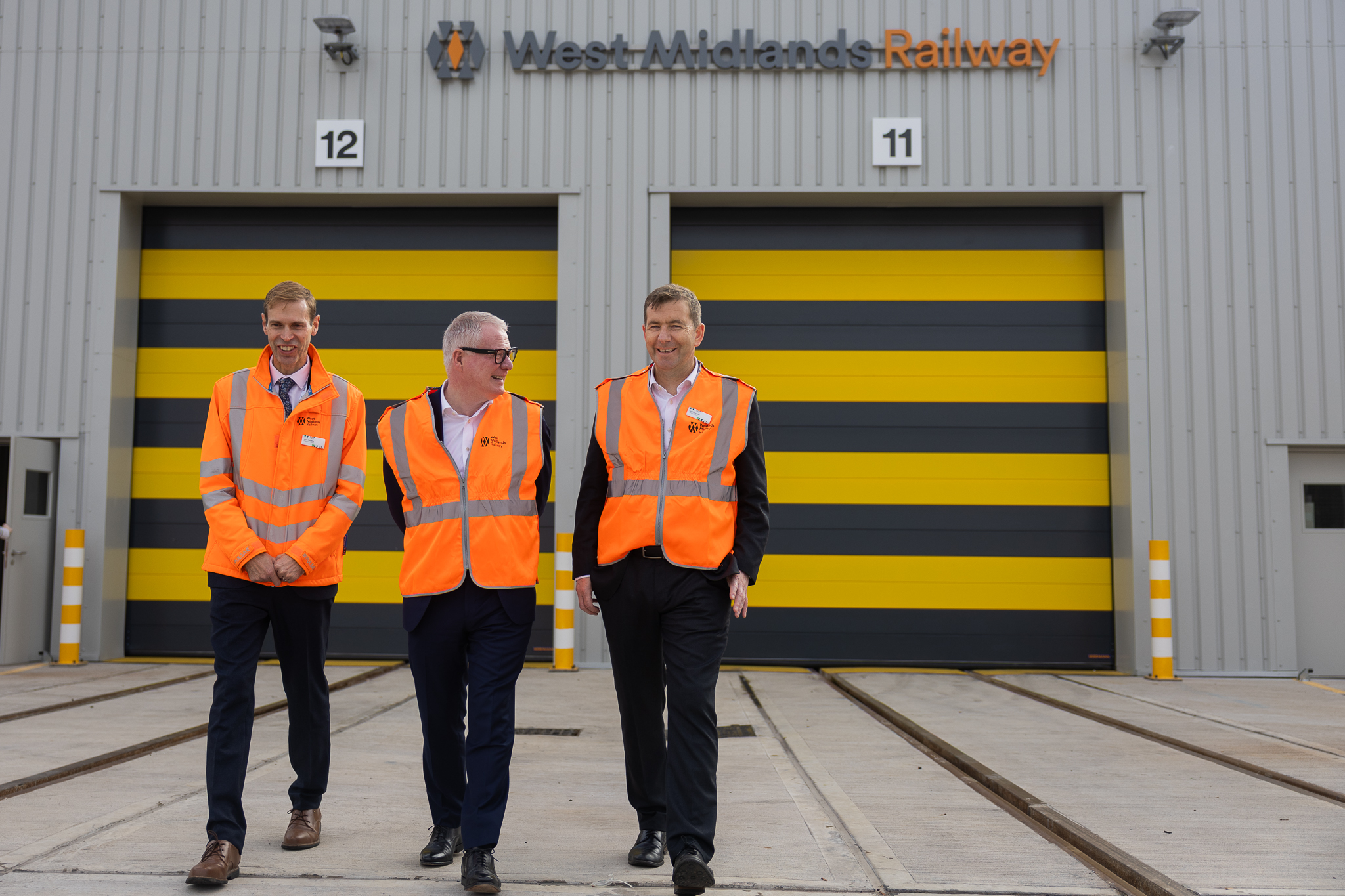 Three men, in orange hi-vis vests walking in front of large metal warehouse building which fills rest of the picture. There are two black and yellow striped garage doors and a sign above says West Midlands Railway