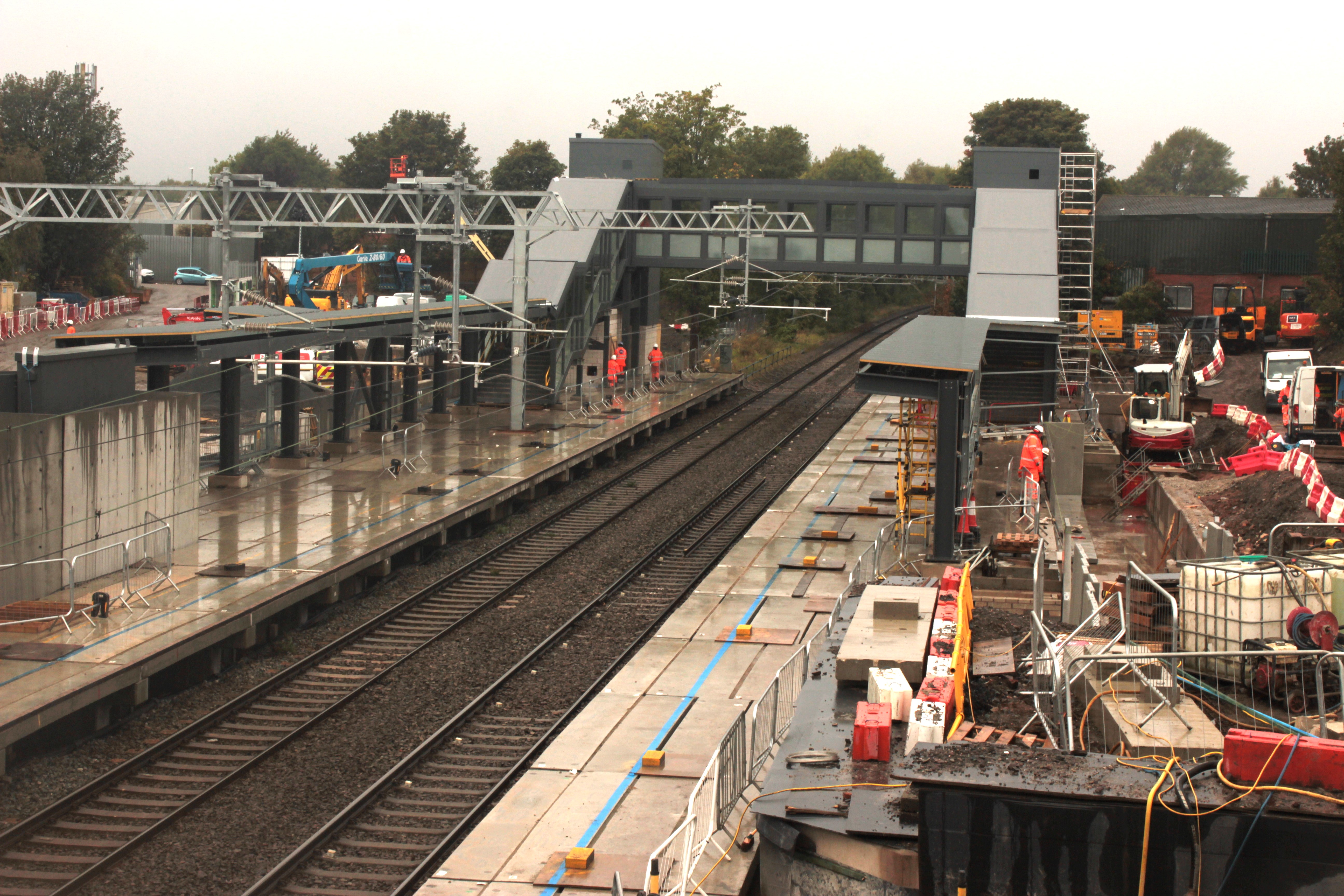 Looking down on a railway station construction site. The tracks and two platforms run through the middle. There are canopies on the platforms and at the far end a footbridge over the track and covered stairways leading up to it. It has been raining and there are orange clad construction workers and building materials scattered around the site.