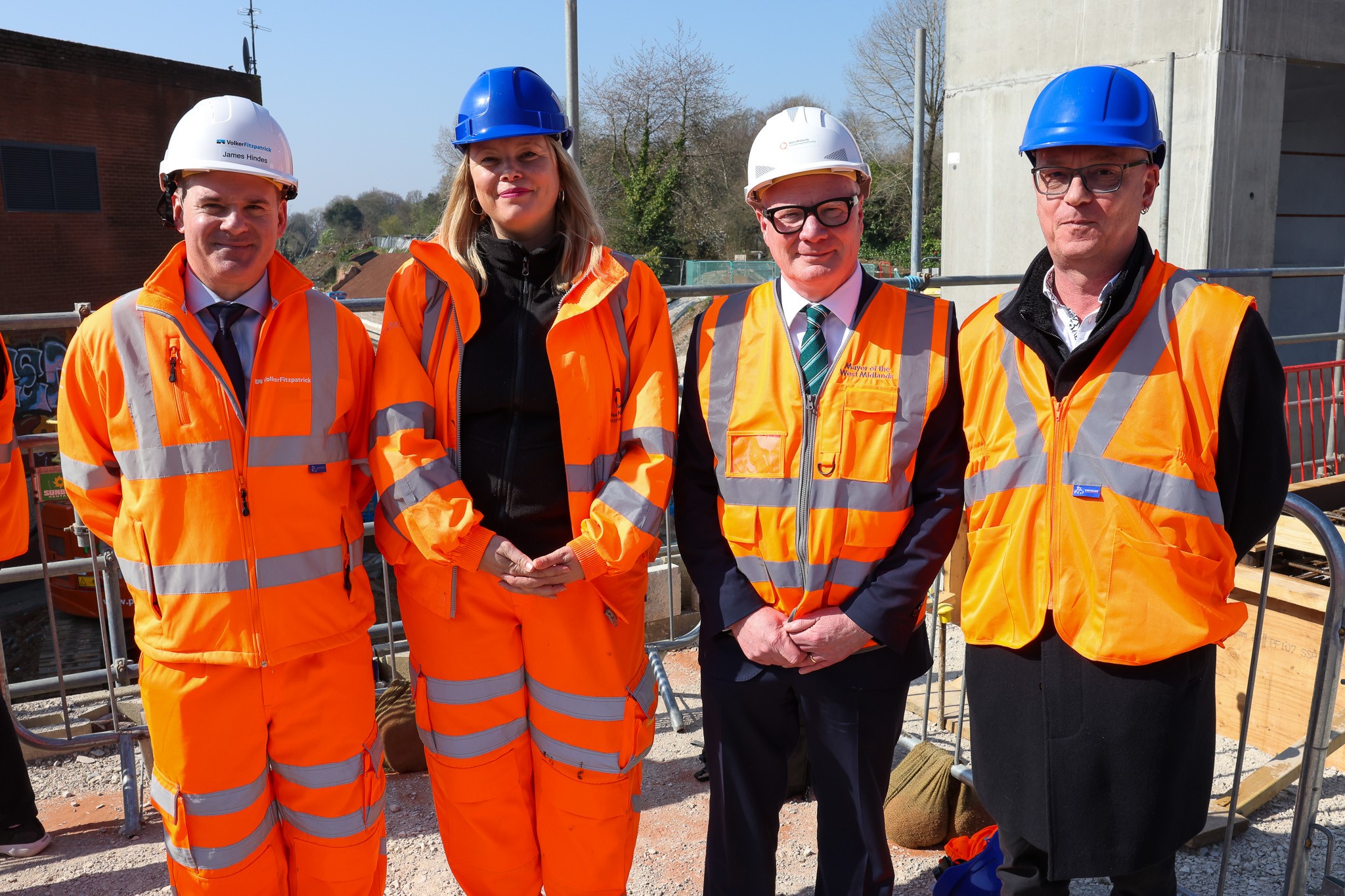 Line up of four VIPs in hard hats and high visibilty orange jackets with the Kings Heath construction site behind - including concrete tower and scaffold poles