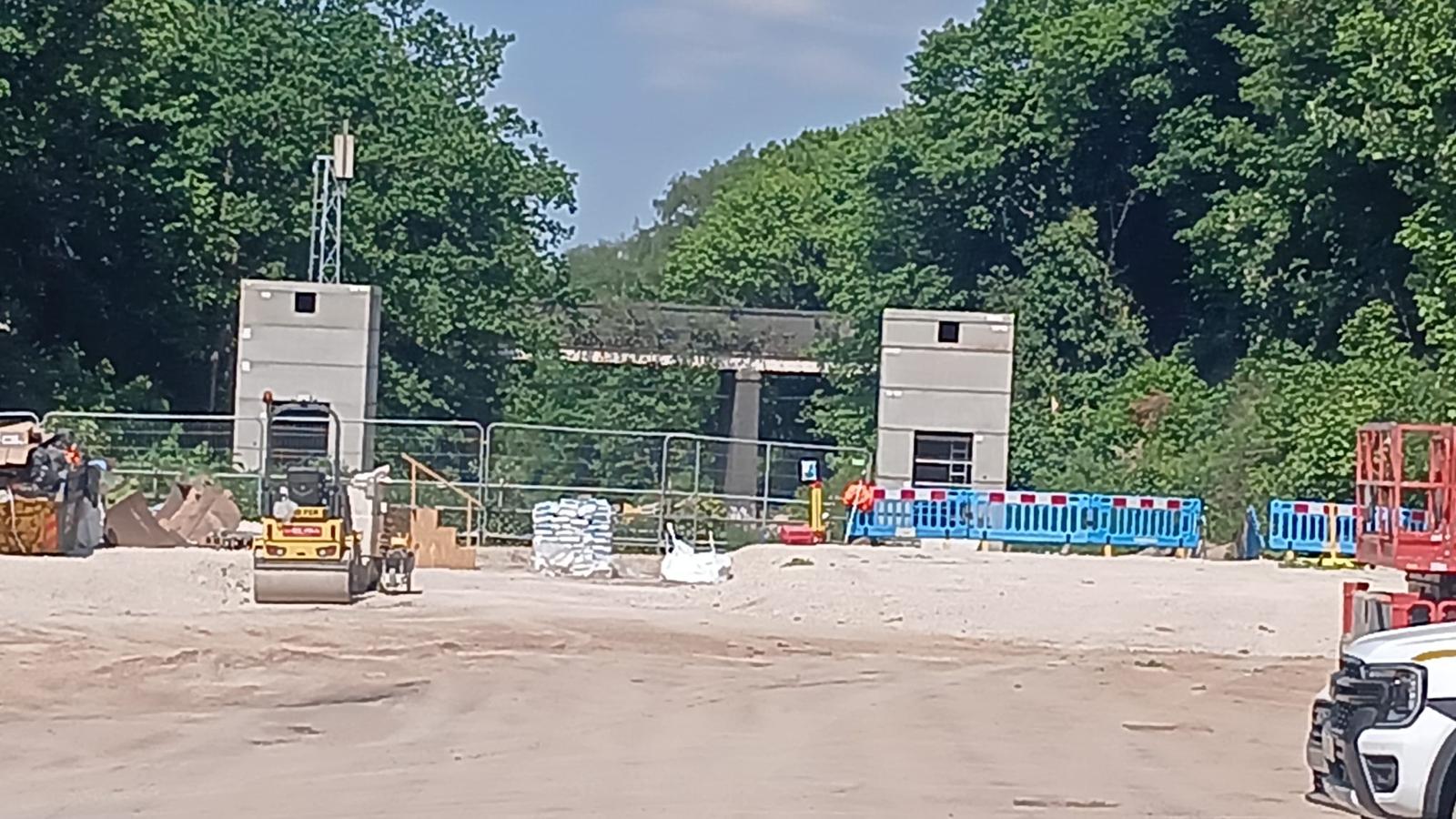 Construction site with, behind fencing, the twin towers of the lift shafts on either side of the frame. Behind are green trees and a road bridge