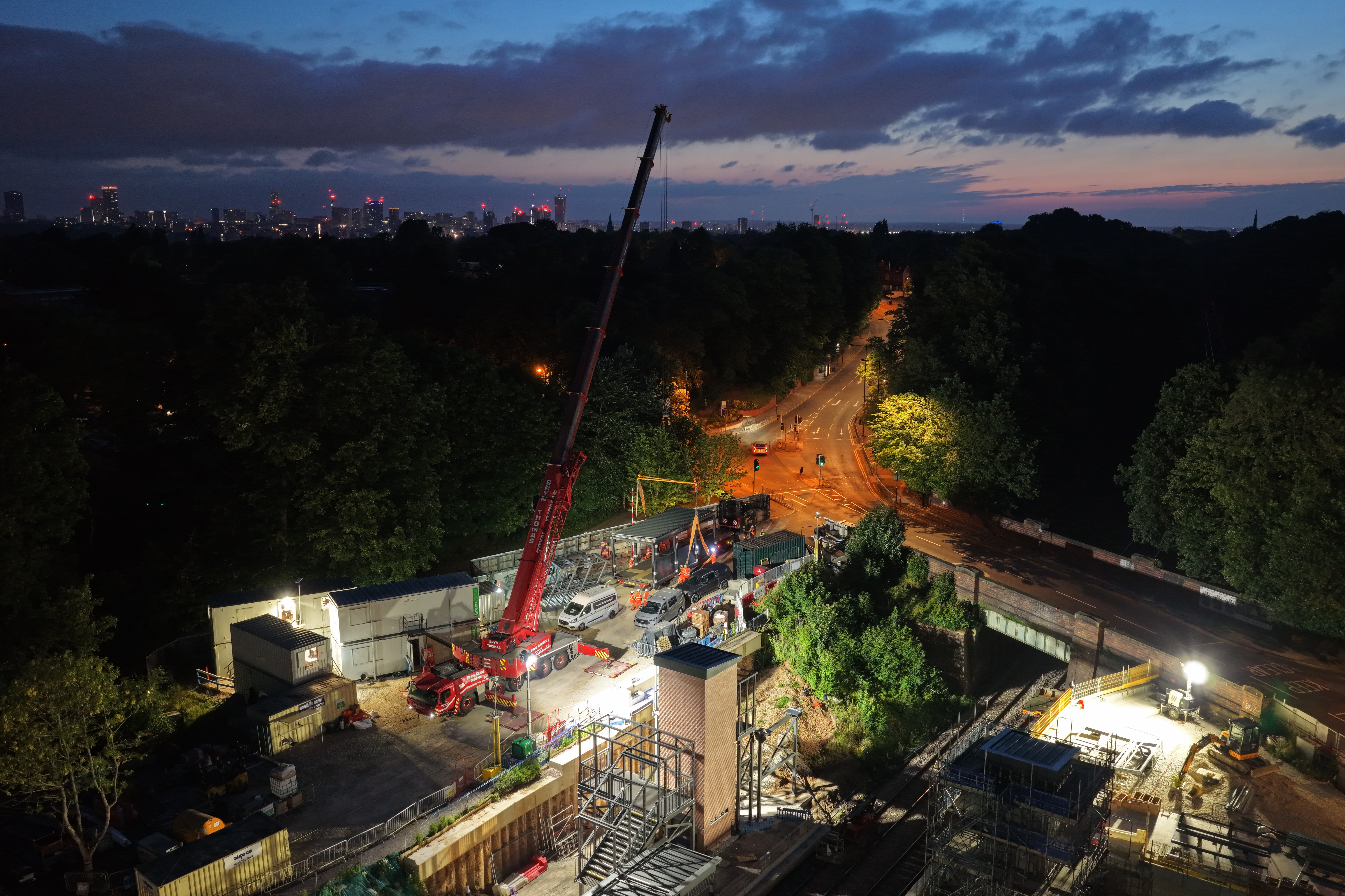 From above overlooking the site - it's night time, but in a spotlit area the orange arm of the crane rises about the railway station with the box bridge dangling below