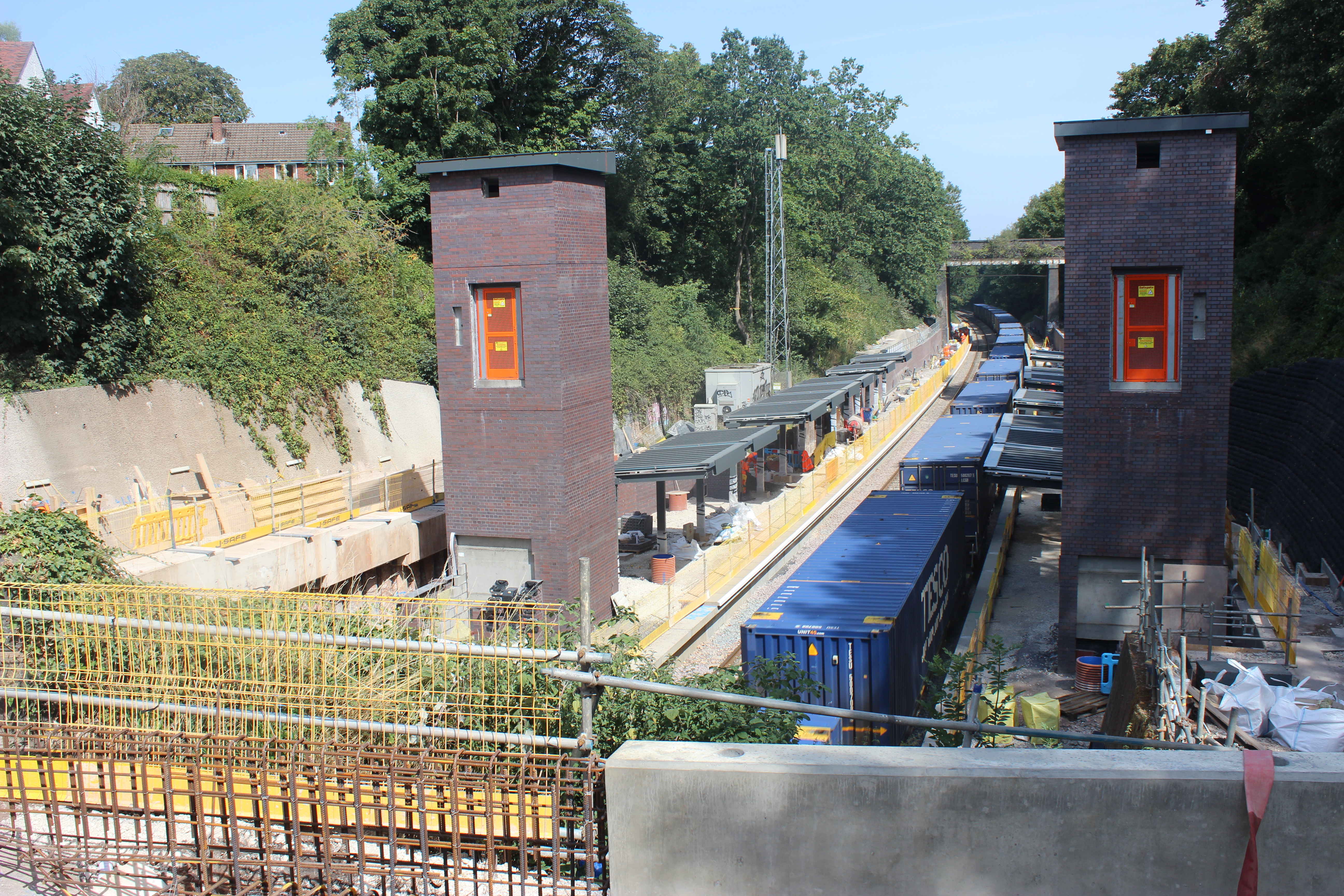 Photo Overlooking a railway station from one side - two platforms stretch off into distance, brick-clad towers which will be passenger lift shafts stand either side . A train with blue box carriages is running along the track.